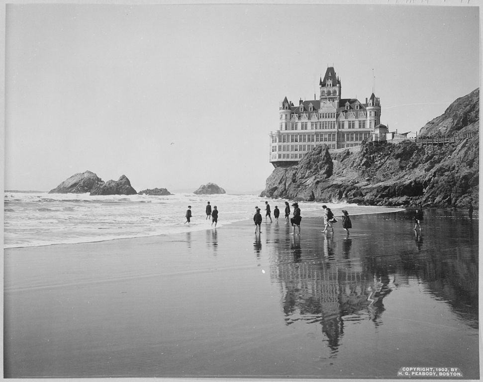 Una imagen en blanco y negro de un hotel en un acantilado junto al mar, en la playa en primer plano pasean personas.