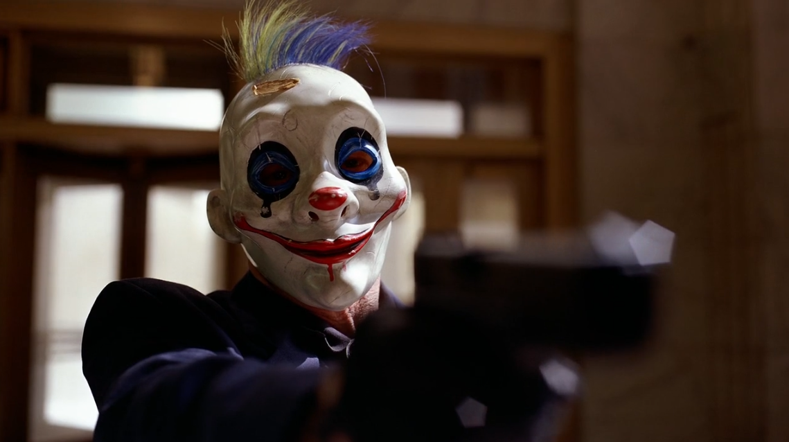 Un atracador con máscara de payaso sonriente con el pelo verde y azul y una chaqueta azul, apunta a la cámara con su pistola.