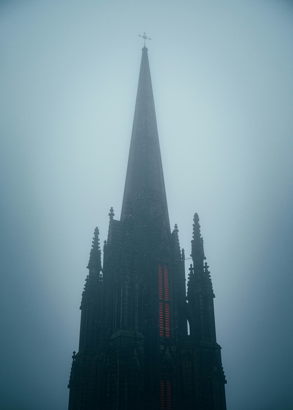 Una torre puntiaguda de estilo gótico con una ventana iluminada en rojo y una cruz encima, entre la niebla.