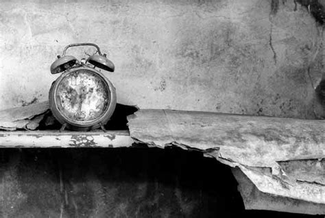 En blanco y negro se ve un reloj despertador antiguo sucio sobre una repisa, a su lado hay fragmentos del techo o la pared.